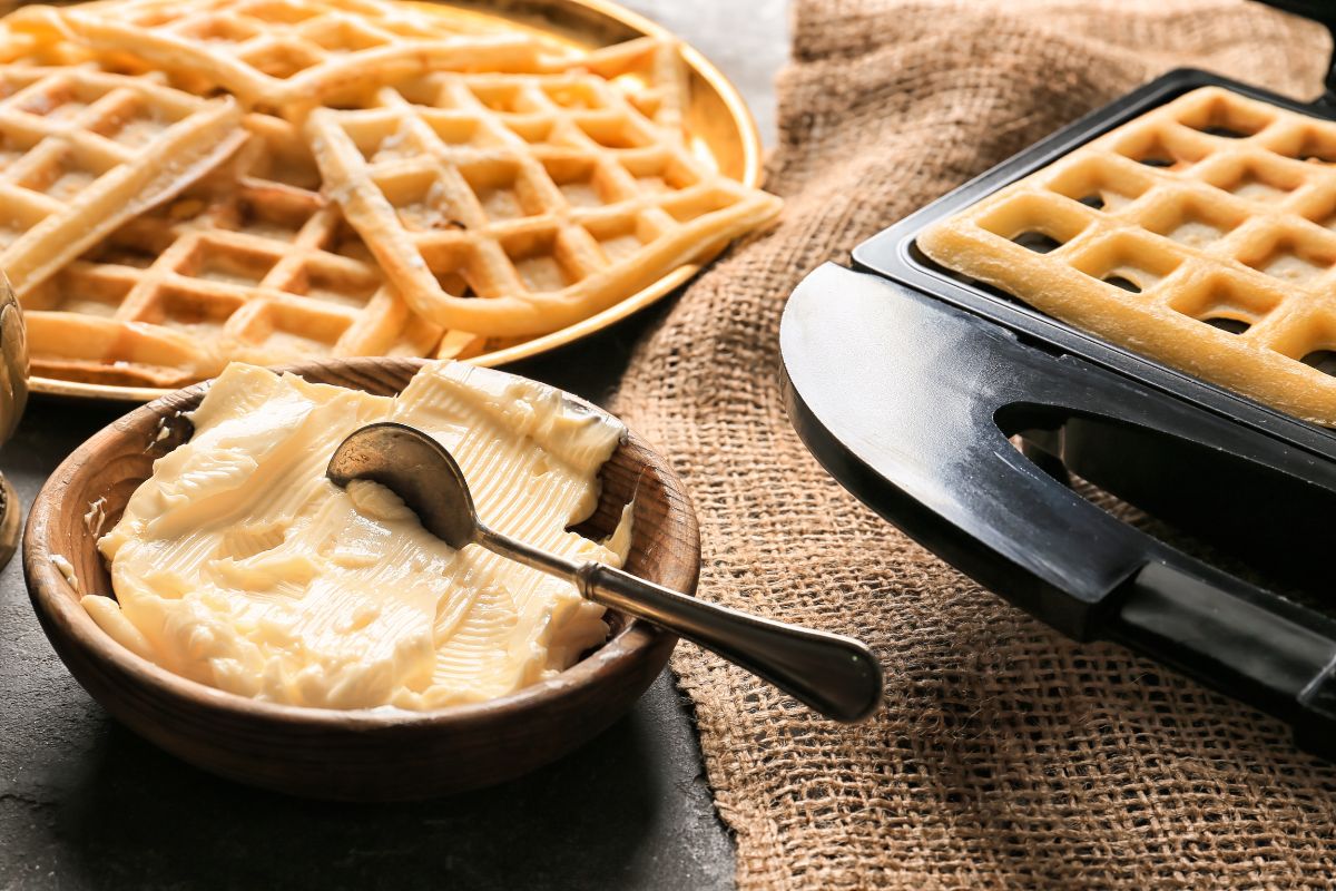 Waffles in a waffle maker, a plate of waffles, and a bowl of butter